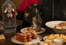 Assassin’s Creed: Xepa terá cardápio especial da franquia Imagem de destaque mostrando o personagem icônico posando em frente a uma mesa repleta de pratos temáticos do evento oficial de Assassin’s Creed no Restaurante Xepa.