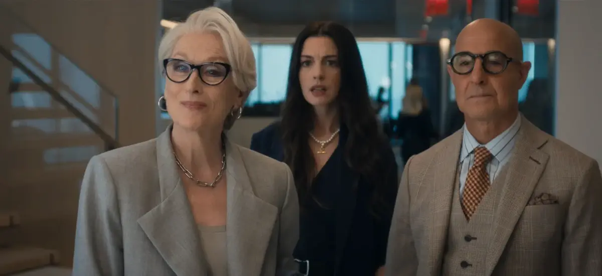 Meryl Streep, Anne Hathaway e Stanley Tucci lado a lado em ambiente corporativo moderno com escada ao fundo em cena do O Diabo Veste Prada 2