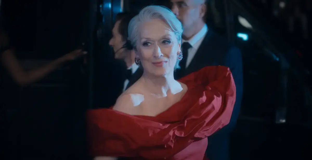 Streep como Miranda Priestly em vestido vermelho dramático com ombro exposto em evento noturno no filme O Diabo Veste Prada 2.