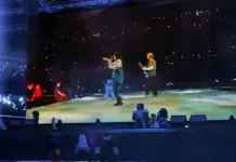 Avatares do Roblox assistem à apresentação de Billie Eilish no The Block, com telão exibindo a cantora no palco durante o evento gratuito de maio de 2026